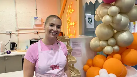 BBC A woman in a pink t-shirt and apron smiles at the camera, in front of a small kitchen in a marketplace. A large display of gold, orange and light pink balloons are next to her.